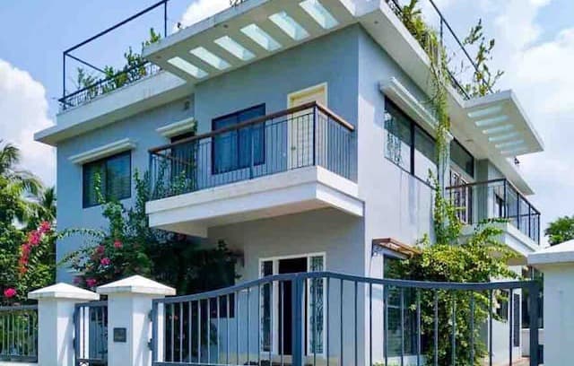 Two-storey blue-grey house with white trim, a slatted white pergola shading the roof terrace, a first-floor balcony with dark railings and a timber top rail, and bougainvillea in flower beside the white-pillared boundary wall and metal gate