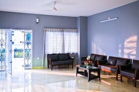 Ground-floor sitting room with blue-grey walls, a glossy cream tiled floor, dark wooden sofa sets around a wooden coffee table with fresh flowers, a ceiling fan, and an ornate white metal grille front door standing open