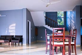 Open-plan dining area with red-brown wooden table and chairs, a ceiling fan, blue-grey walls and glossy tiled floor, beside an internal staircase with a dark timber handrail and a tall grilled window looking onto greenery