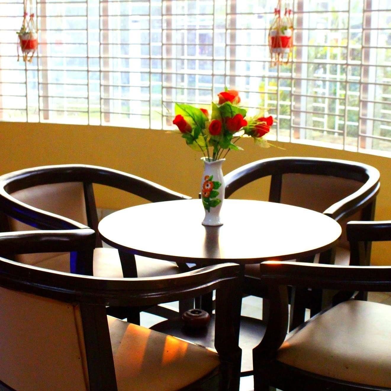 Sunlit sitting corner with a round table, dark wood armchairs, a vase of red roses, and hanging planters against a grilled window