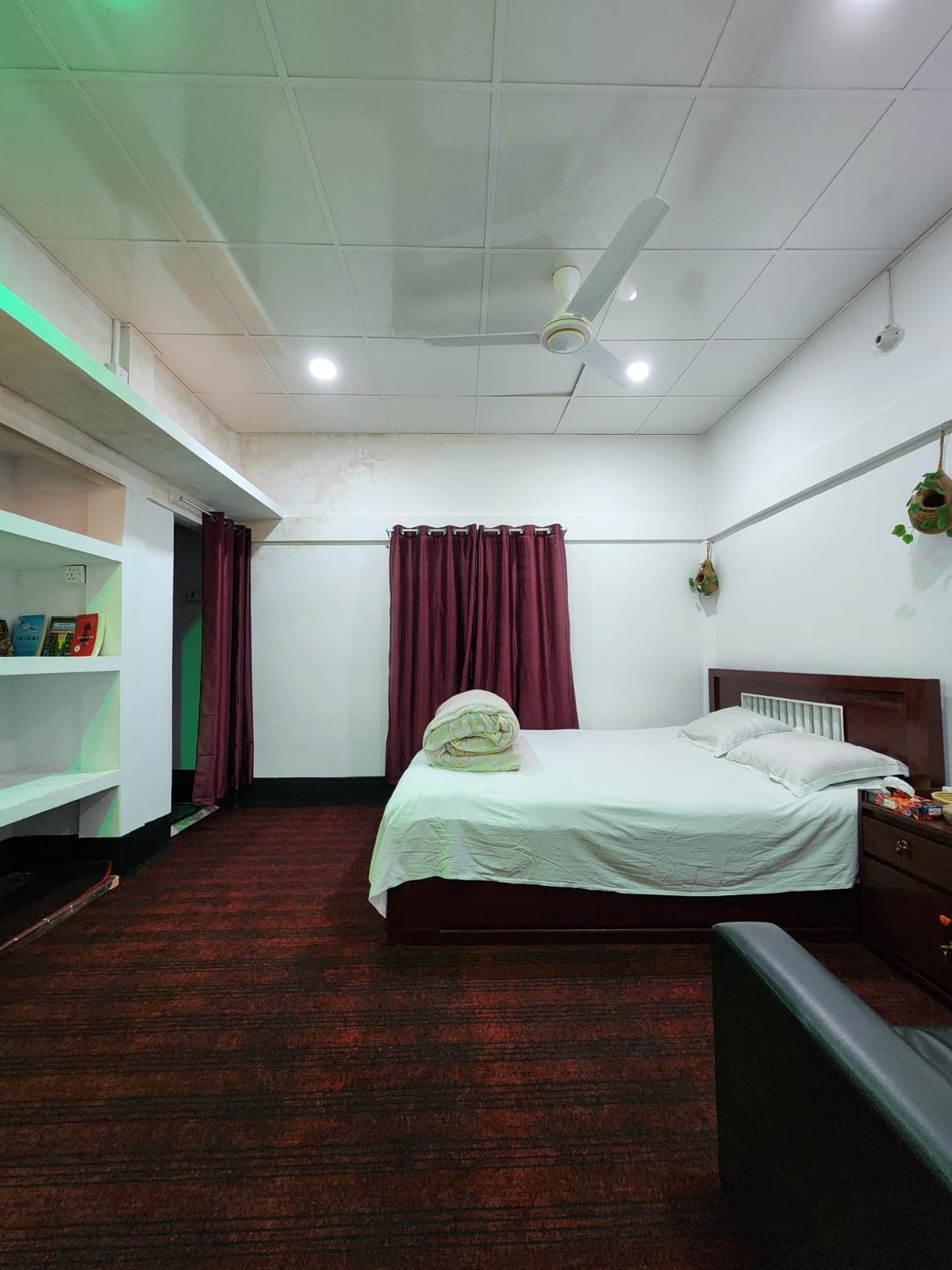 Guest room with maroon curtains, a small bookshelf stocked with books, and woven hanging planters on the wall