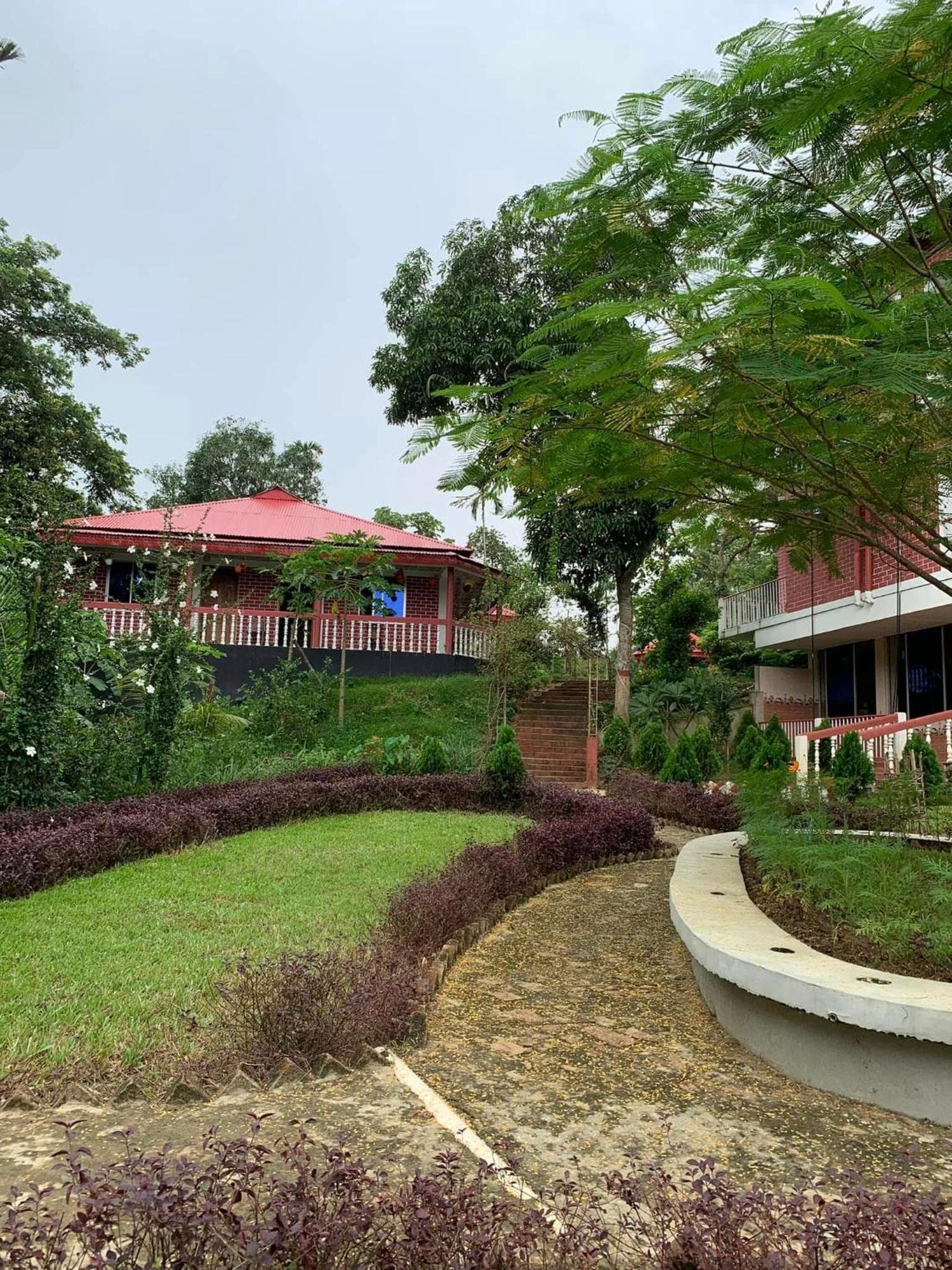 Landscaped garden with a curving paved path and burgundy ornamental hedge borders around a lawn, brick steps rising past conifer shrubs to a red tin-roofed brick cottage with a red-and-white balustrade verandah, and a red-brick guest building with a glazed ground floor at the right edge