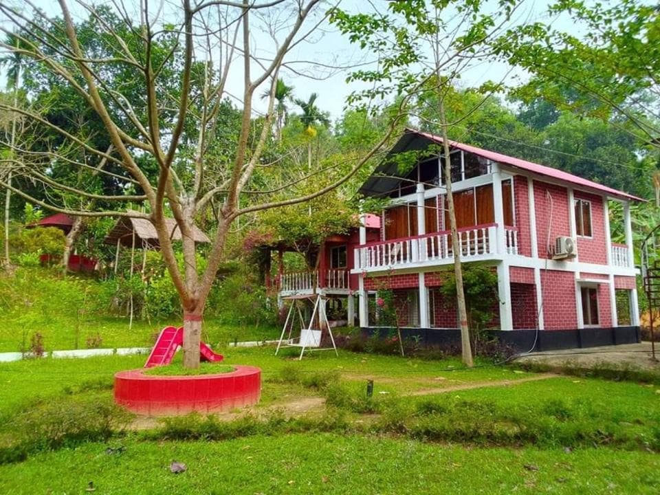 Two-storey pink-brick guest building with white columns and white balcony railings under a tin roof, with a red children's slide, a white A-frame swing seat and a red circular tree-ring planter on the rough grass in front