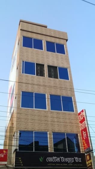 Daytime street view of the tan-tiled Tower Inn building with its Bangla signboard "হোটেল টাওয়ার ইন" (Hotel Tower Inn) above the ground-floor shop